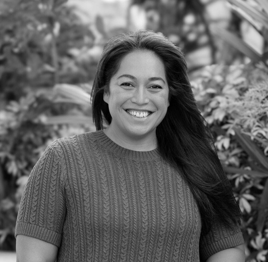 Smiling woman with long hair wearing a knitted sweater, surrounded by greenery, representing the Integrated Wellness Clinic's Sunshine Coast Naturopath team.