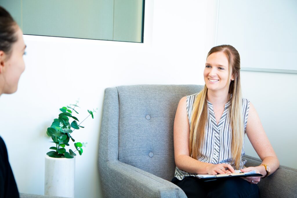 Smiling female practitioner in a striped blouse sitting in a modern therapy room, engaging with a client, with a potted plant in the foreground, reflecting the Integrated Wellness Clinic's focus on holistic health and wellness.