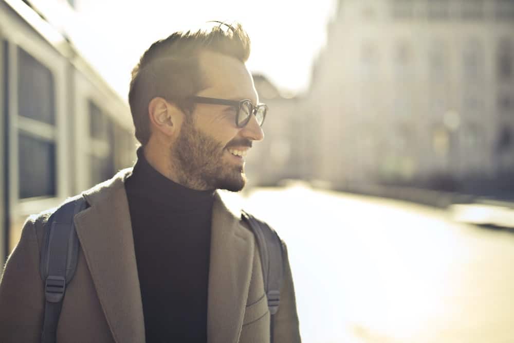 Smiling man in a stylish coat and glasses, walking outdoors in a bright urban setting, representing confidence and well-being, aligned with mental health and holistic wellness themes.