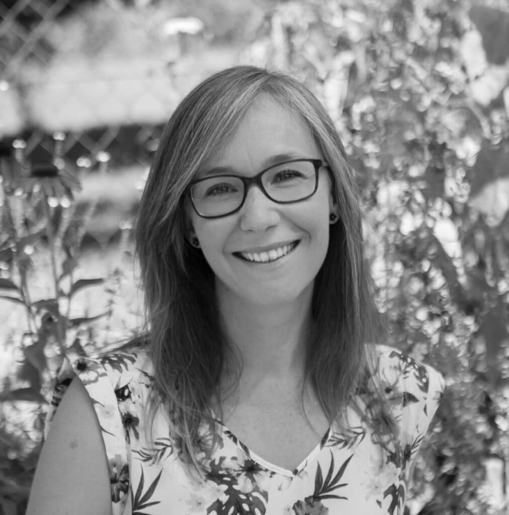 Alyson Maier, smiling, wearing glasses, in a floral top, surrounded by greenery, representing expertise in anxiety, mood, children's health, and hormonal imbalance at Integrated Wellness Clinic.