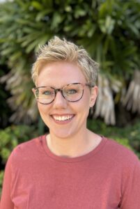 Smiling woman with short blonde hair and glasses, wearing a red shirt, surrounded by greenery, representing Lauren Baker, a Registered Clinical Counsellor specializing in marriage and family therapy at Integrated Wellness Clinic.
