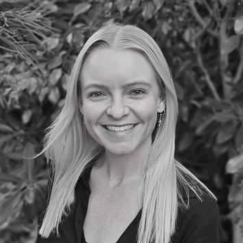 Smiling naturopath with long blonde hair, wearing a black top, surrounded by greenery, representing the Integrated Wellness Clinic's holistic health approach.